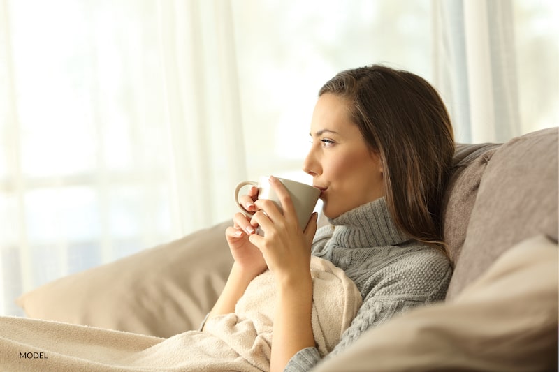 Woman resting under a blanket, drinking a cup of tea. 