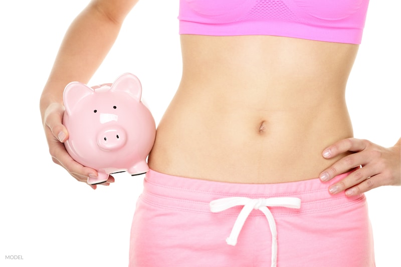 Women with slim abdomen holding a piggy bank in pink workout clothes.